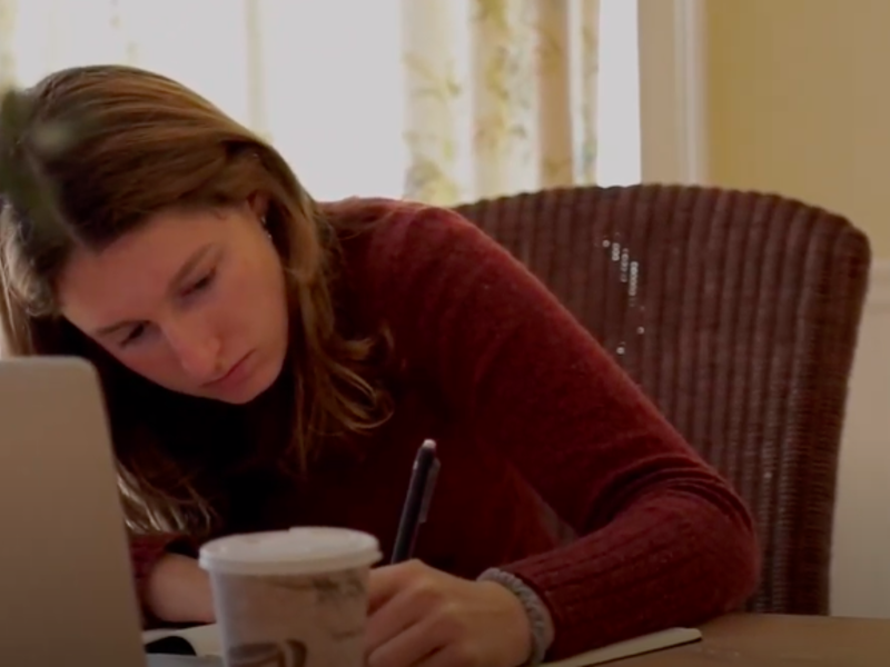 Young woman writing in a notebook, sitting in front of her laptop.