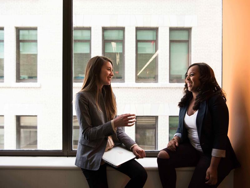 Two women in business attire chat on a window ledge.